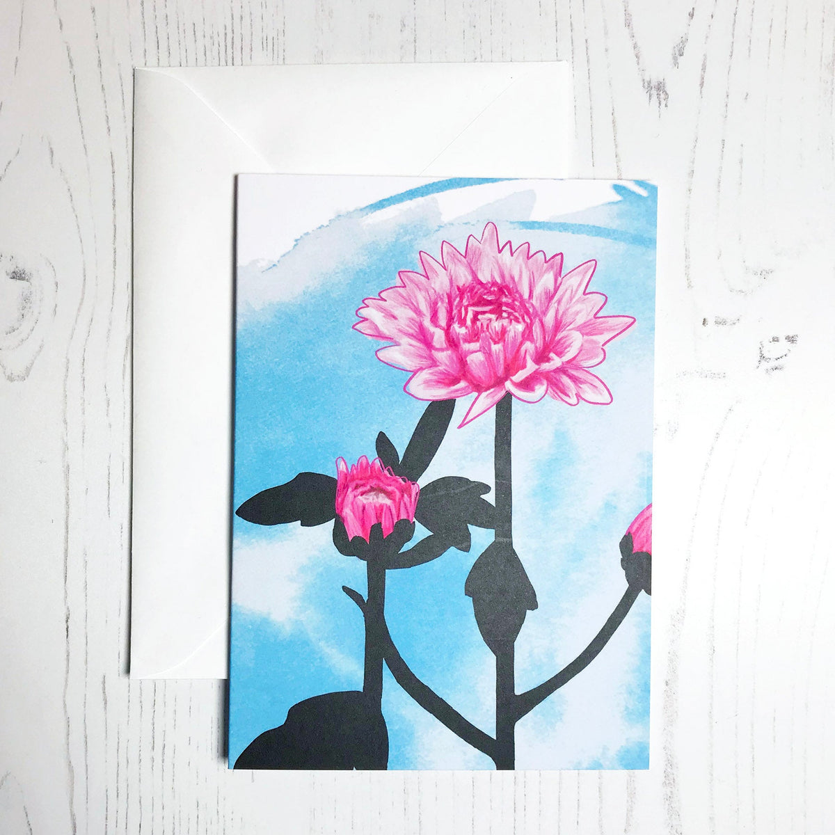 An A6 greetings card showing an illustration of pink chrysanthemums against an inky blue wash background. The flowers are in colour with the plant leaves silhouetted in black.