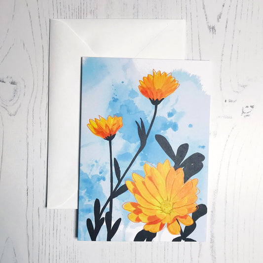 An A6 greetings card showing an illustration of marigolds against an inky blue wash background. The marigolds are warm yellows and oranges  with the plant leaves silhouetted in black.