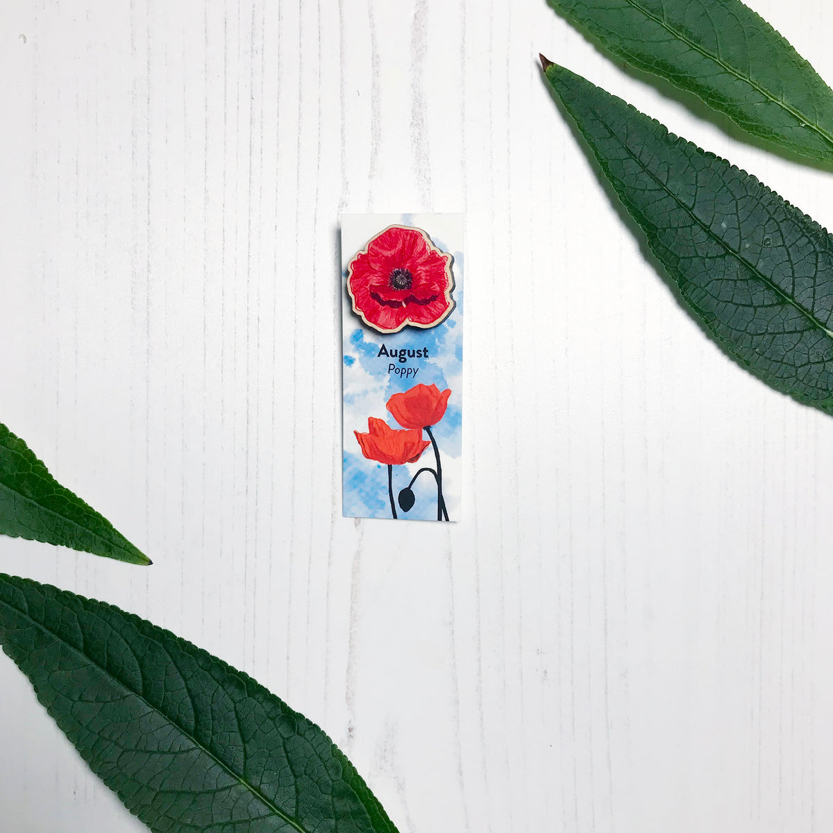 A 25mm mini wooden lapel pin of a red poppy flower mounted on an illustrated backing card. The pin is made from an original illustration and is loose and expressive in style. The backing card says August Poppy and has an illustration of red poppies with stems silhouetted in black against an inky blue background.