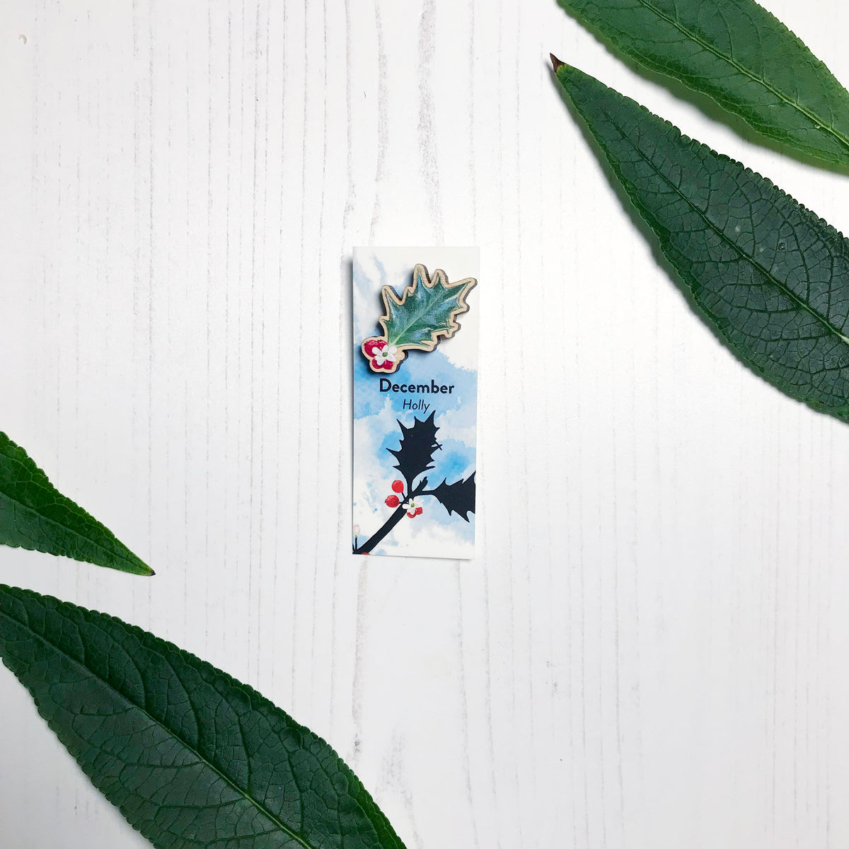 A 25mm mini wooden lapel pin of a holly leaf with red berries mounted on an illustrated backing card. The pin is made from an original illustration and is loose and expressive in style. The backing card says December Holly and has an illustration of red berries with stems and foliage silhouetted in black against an inky blue background.