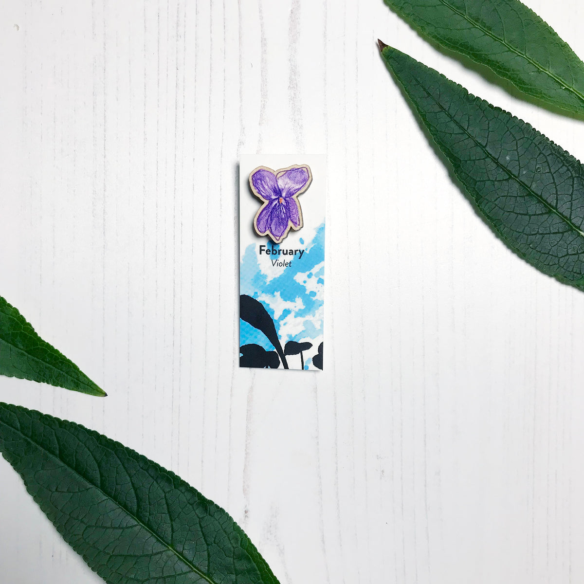 A 25mm mini wooden lapel pin of a purple violet flower mounted on an illustrated backing card. The pin is made from an original illustration and is loose and expressive in style. The backing card says February Violet and has an illustration of violet plant foliage silhouetted in black against an inky blue background.