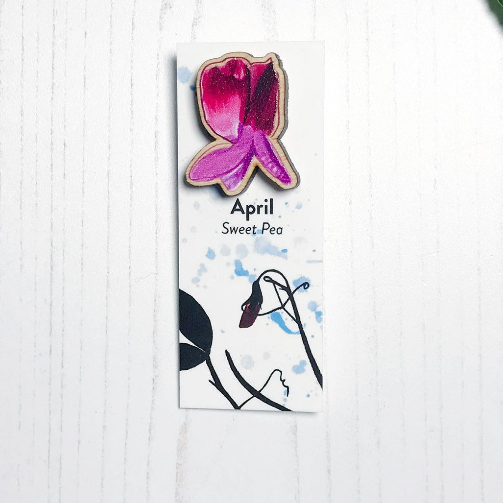 A 25mm mini wooden lapel pin of a pinky purple sweet pea flower mounted on an illustrated backing card. The pin is made from an original illustration and is loose and expressive in style. The backing card says April Sweet Pea and has an illustration of sweet pea foliage silhouetted in black against an inky blue background.