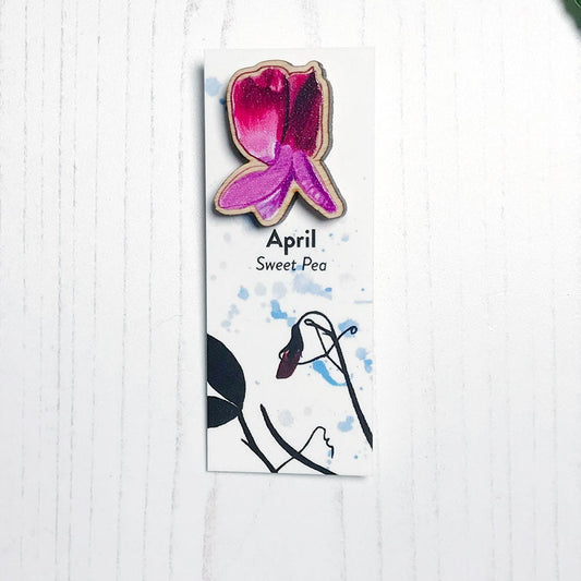 A 25mm mini wooden lapel pin of a pinky purple sweet pea flower mounted on an illustrated backing card. The pin is made from an original illustration and is loose and expressive in style. The backing card says April Sweet Pea and has an illustration of sweet pea foliage silhouetted in black against an inky blue background.