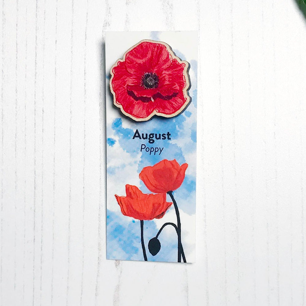 A 25mm mini wooden lapel pin of a red poppy flower mounted on an illustrated backing card. The pin is made from an original illustration and is loose and expressive in style. The backing card says August Poppy and has an illustration of red poppies with stems silhouetted in black against an inky blue background.