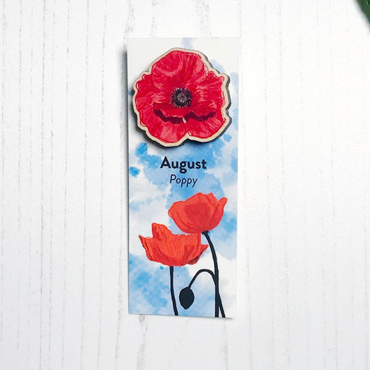 A 25mm mini wooden lapel pin of a red poppy flower mounted on an illustrated backing card. The pin is made from an original illustration and is loose and expressive in style. The backing card says August Poppy and has an illustration of red poppies with stems silhouetted in black against an inky blue background.