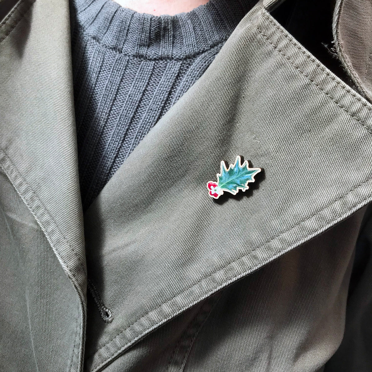 A 25mm mini wooden lapel pin of a holly leaf with red berries worn on the lapel of a jacket. The pin is made from an original illustration and is loose and expressive in style. 