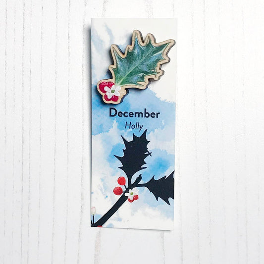 A 25mm mini wooden lapel pin of a holly leaf with red berries mounted on an illustrated backing card. The pin is made from an original illustration and is loose and expressive in style. The backing card says December Holly and has an illustration of red berries with stems and foliage silhouetted in black against an inky blue background.