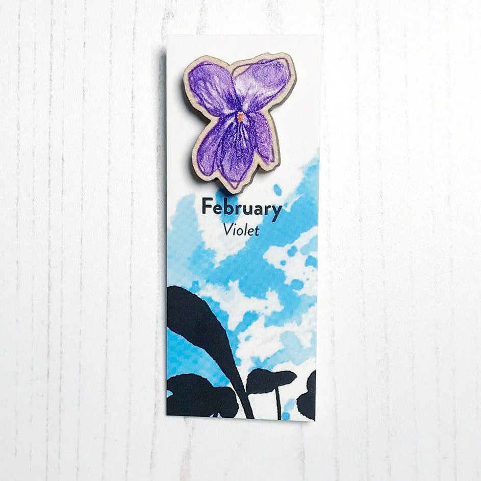 A 25mm mini wooden lapel pin of a purple violet flower mounted on an illustrated backing card. The pin is made from an original illustration and is loose and expressive in style. The backing card says February Violet and has an illustration of violet plant foliage silhouetted in black against an inky blue background.