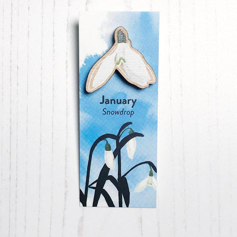 A 25mm mini wooden lapel pin of a snowdrop flower mounted on an illustrated backing card. The pin is made from an original illustration and is loose and expressive in style. The backing card says January Snowdrop and has an illustration of snowdrops on where the flowers are in colour with silhouette leaves against an inky blue background.