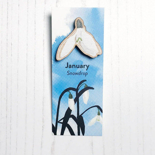 A 25mm mini wooden lapel pin of a snowdrop flower mounted on an illustrated backing card. The pin is made from an original illustration and is loose and expressive in style. The backing card says January Snowdrop and has an illustration of snowdrops on where the flowers are in colour with silhouette leaves against an inky blue background.
