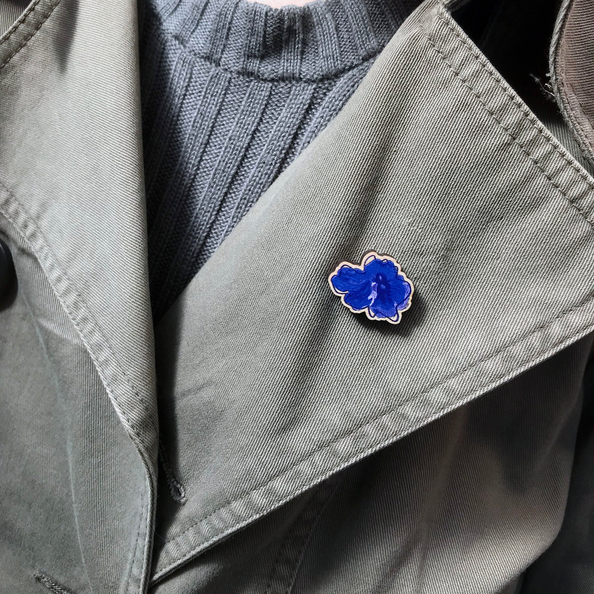 A 25mm mini wooden lapel pin of a blue delphinium flower worn on the lapel of a jacket. The pin is made from an original illustration and is loose and expressive in style. 
