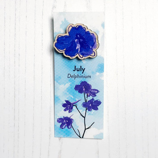 A 25mm mini wooden lapel pin of a blue delphinium flower mounted on an illustrated backing card. The pin is made from an original illustration and is loose and expressive in style. The backing card says July Delphinium and has an illustration of blue delphinium with foliage and stems silhouetted in black against an inky blue background.