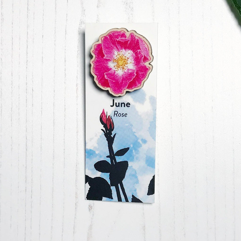 A 25mm mini wooden lapel pin of a pink dog rose flower mounted on an illustrated backing card. The pin is made from an original illustration and is loose and expressive in style. The backing card says June Rose and has an illustration of rose buds with foliage and stems silhouetted in black against an inky blue background.