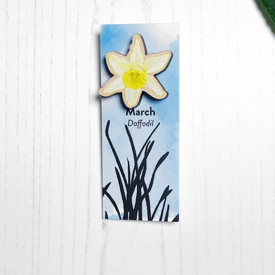 A 25mm mini wooden lapel pin of a yellow daffodil flower mounted on an illustrated backing card. The pin is made from an original illustration and is loose and expressive in style. The backing card says March Daffodil and has an illustration of daffodil plant foliage silhouetted in black against an inky blue background.