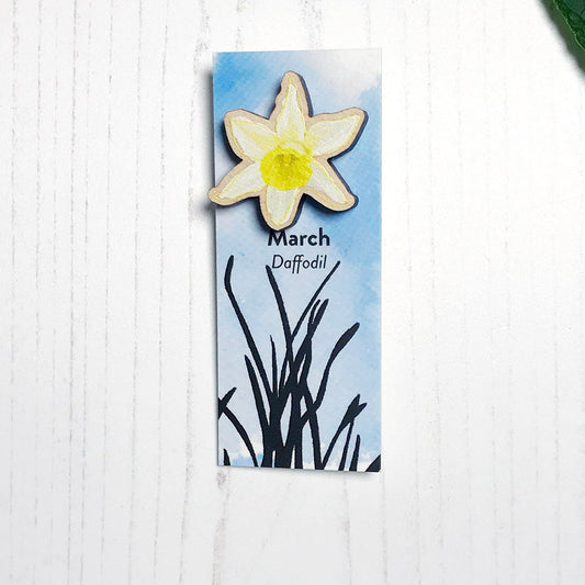 A 25mm mini wooden lapel pin of a yellow daffodil flower mounted on an illustrated backing card. The pin is made from an original illustration and is loose and expressive in style. The backing card says March Daffodil and has an illustration of daffodil plant foliage silhouetted in black against an inky blue background.