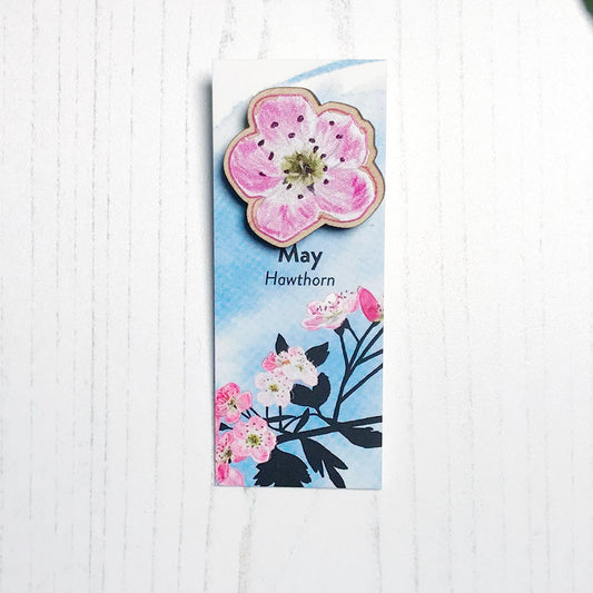 A 25mm mini wooden lapel pin of a pinky white hawthorn blossom mounted on an illustrated backing card. The pin is made from an original illustration and is loose and expressive in style. The backing card says May Hawthorn and has an illustration of Hawthorn blossoms with foliage silhouetted in black against an inky blue background.