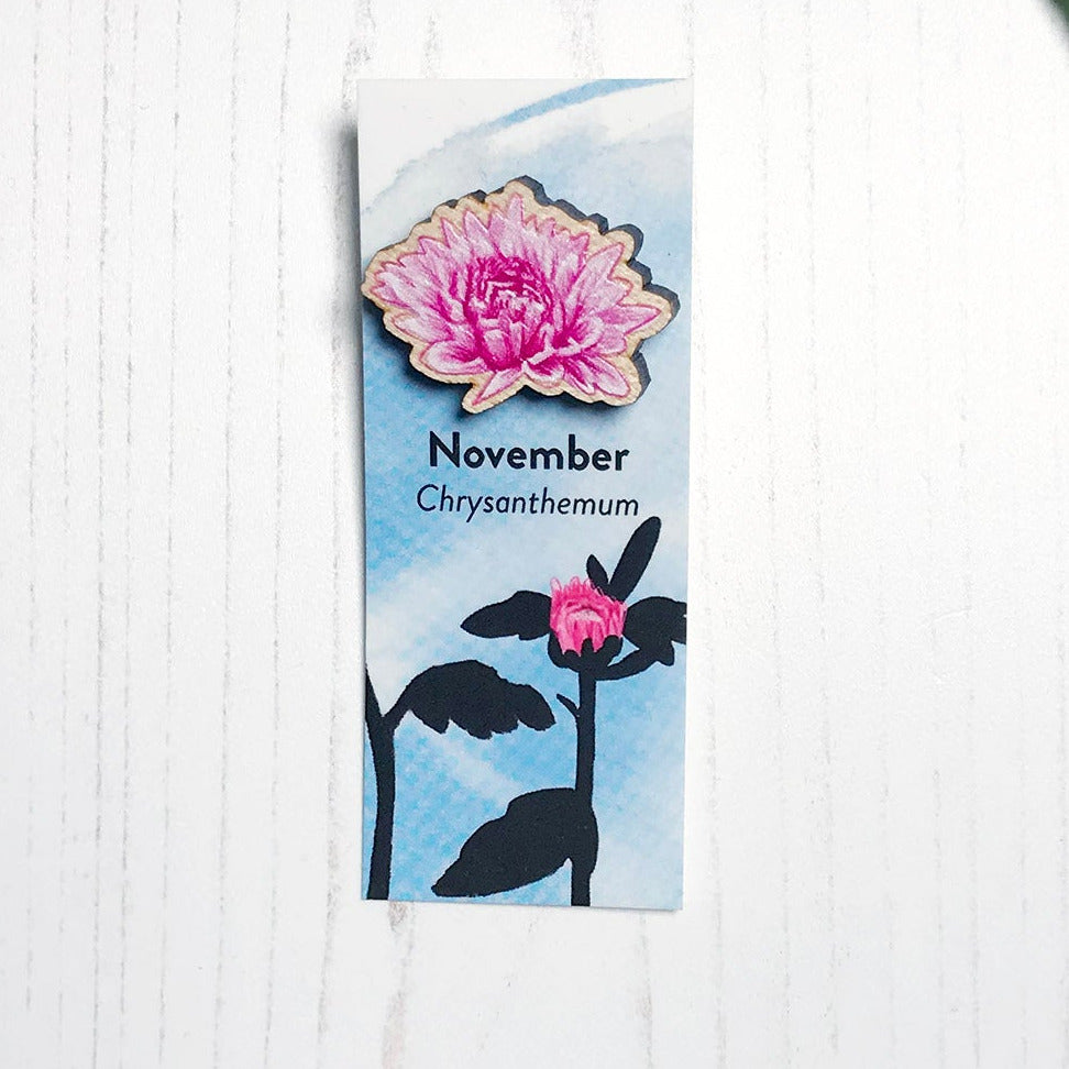 A 25mm mini wooden lapel pin of a pink chrysanthemum flower mounted on an illustrated backing card. The pin is made from an original illustration and is loose and expressive in style. The backing card says November Chrysanthemum and has an illustration of pink chrysanthemum bud with stems and foliage silhouetted in black against an inky blue background.