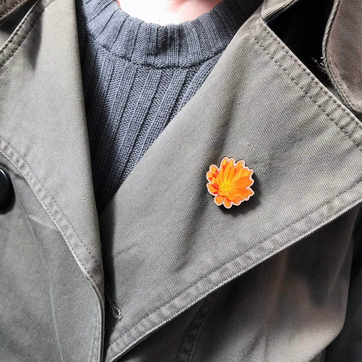 A 25mm mini wooden lapel pin of a orange yellow marigold flower worn on the lapel of a jacket. The pin is made from an original illustration and is loose and expressive in style. 