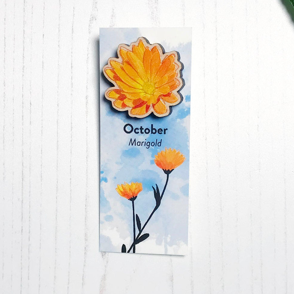 A 25mm mini wooden lapel pin of a orange yellow marigold flower mounted on an illustrated backing card. The pin is made from an original illustration and is loose and expressive in style. The backing card says October Marigold and has an illustration of yellow orange marigolds with stems and foliage silhouetted in black against an inky blue background.