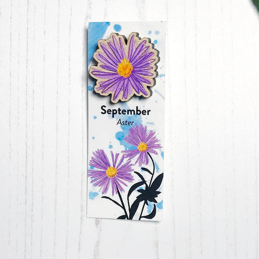 A 25mm mini wooden lapel pin of a purple aster flower mounted on an illustrated backing card. The pin is made from an original illustration and is loose and expressive in style. The backing card says September Aster and has an illustration of purple asters with stems and foliage silhouetted in black against an inky blue background.
