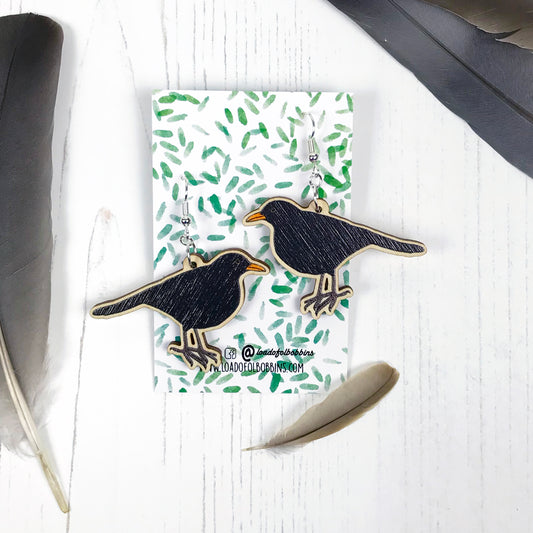 A pair of laser cut wooden dangle earrings showing an illustration by Loadofolbobbins of a male blackbird. The earrings are mounted on a backing card with a repeat abstract watercolour leaf pattern.