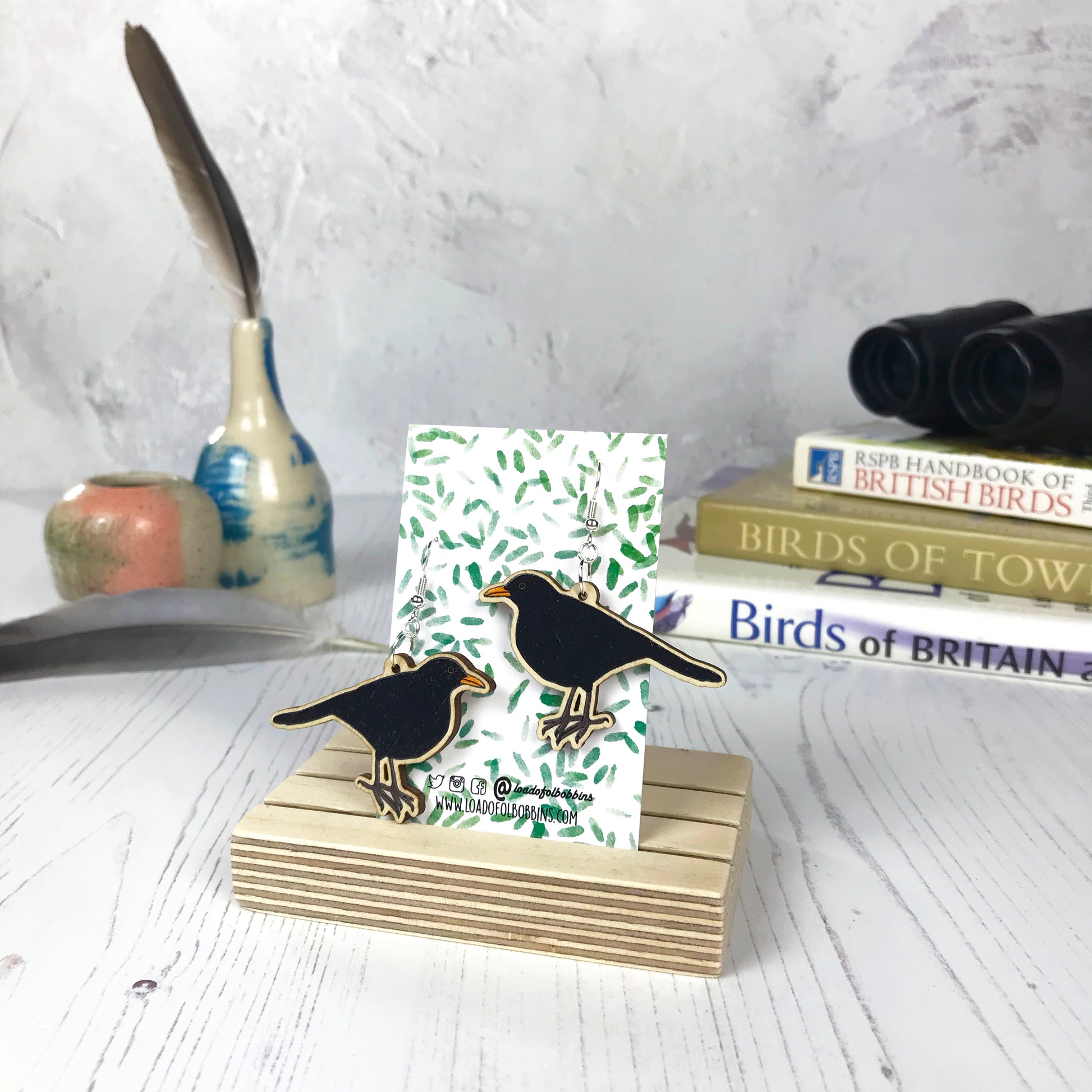 A pair of laser cut wooden dangle earrings showing an illustration of a male blackbird by loadofolbobbins. The earrings are mounted on a backing card with a repeat abstract watercolour leaf pattern. They are standing in a wooden stand and in the background you can see a pile of bird watching books and a pair of binoculars.