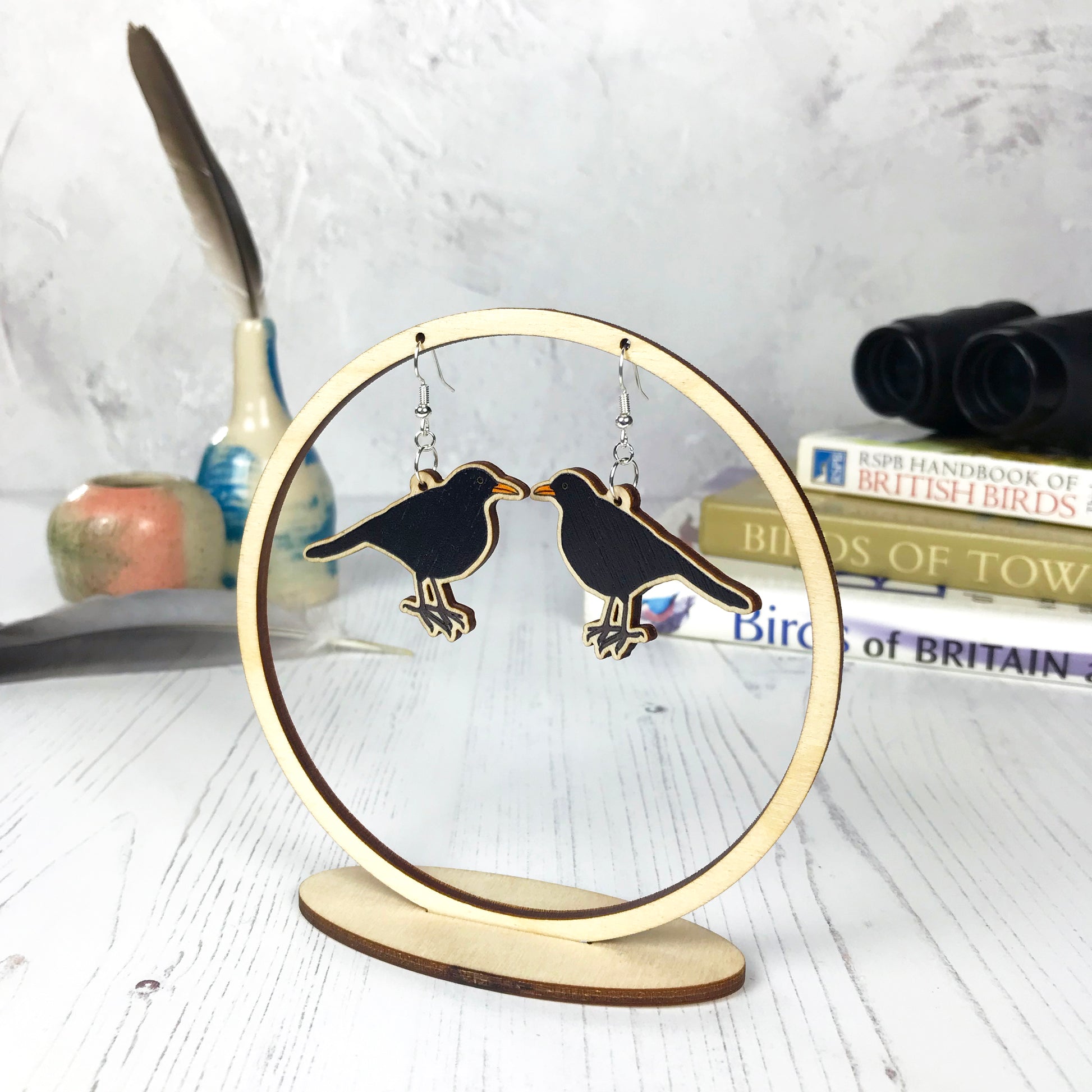 A pair of laser cut wooden dangle earrings showing an illustration of a male blackbird by loadofolbobbins. The earrings are hanging from a wooden circular ring display. 