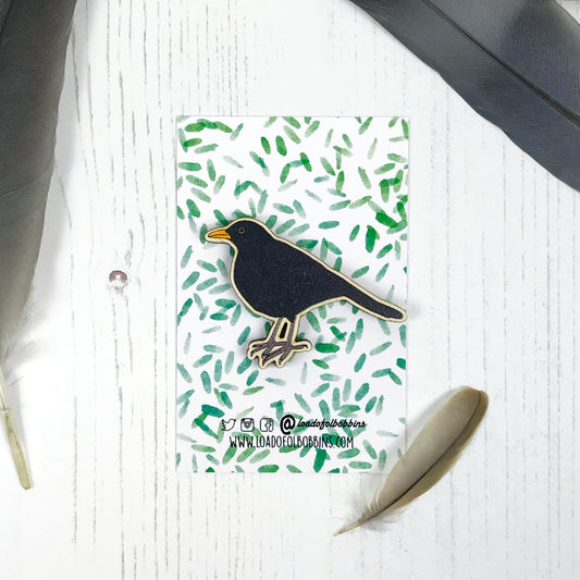 A laser cut wooden brooch showing an illustration by Loadofolbobbins of a male blackbird. The brooch is mounted on a backing card with a repeat abstract watercolour leaf pattern.