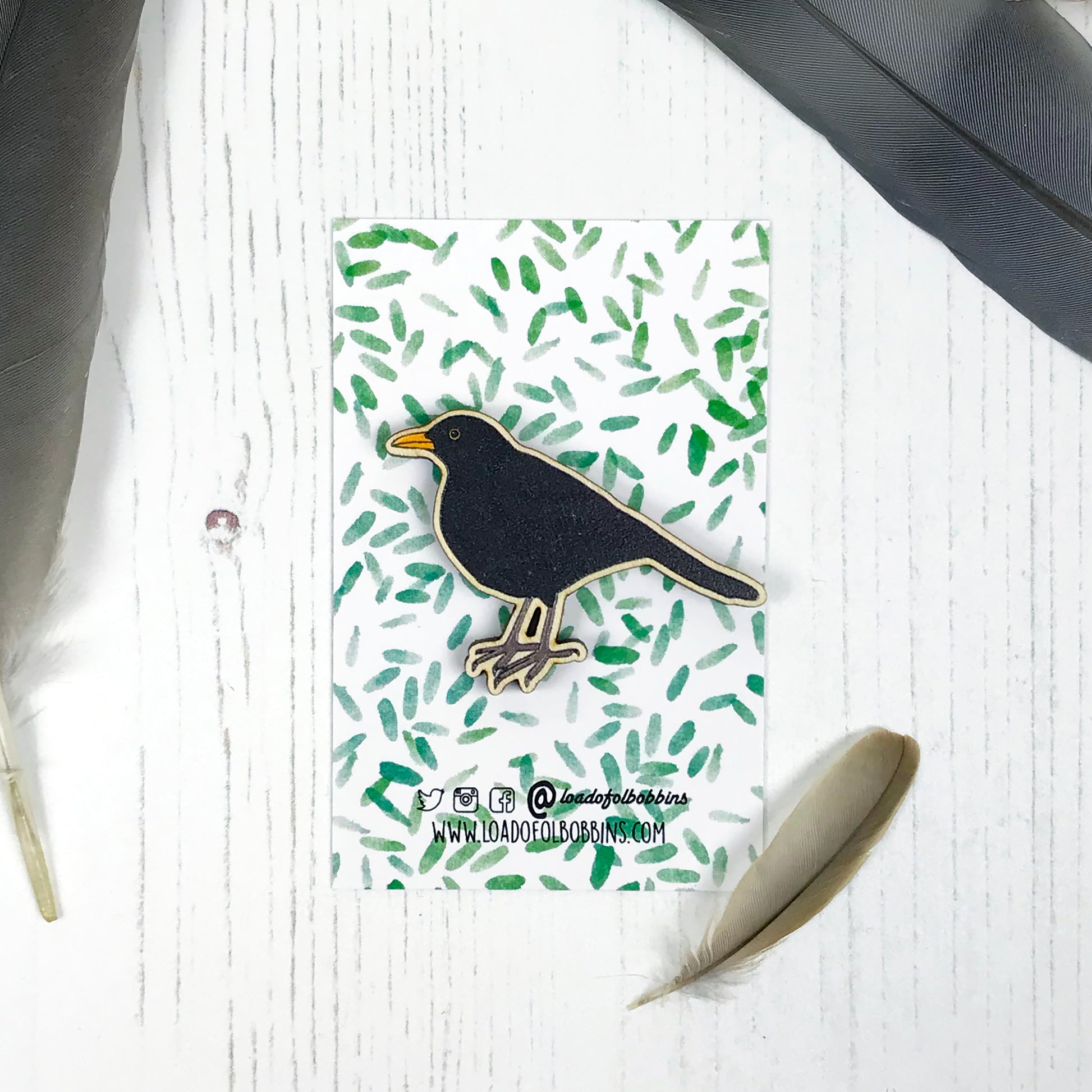 A laser cut wooden brooch showing an illustration by Loadofolbobbins of a male blackbird. The brooch is mounted on a backing card with a repeat abstract watercolour leaf pattern.