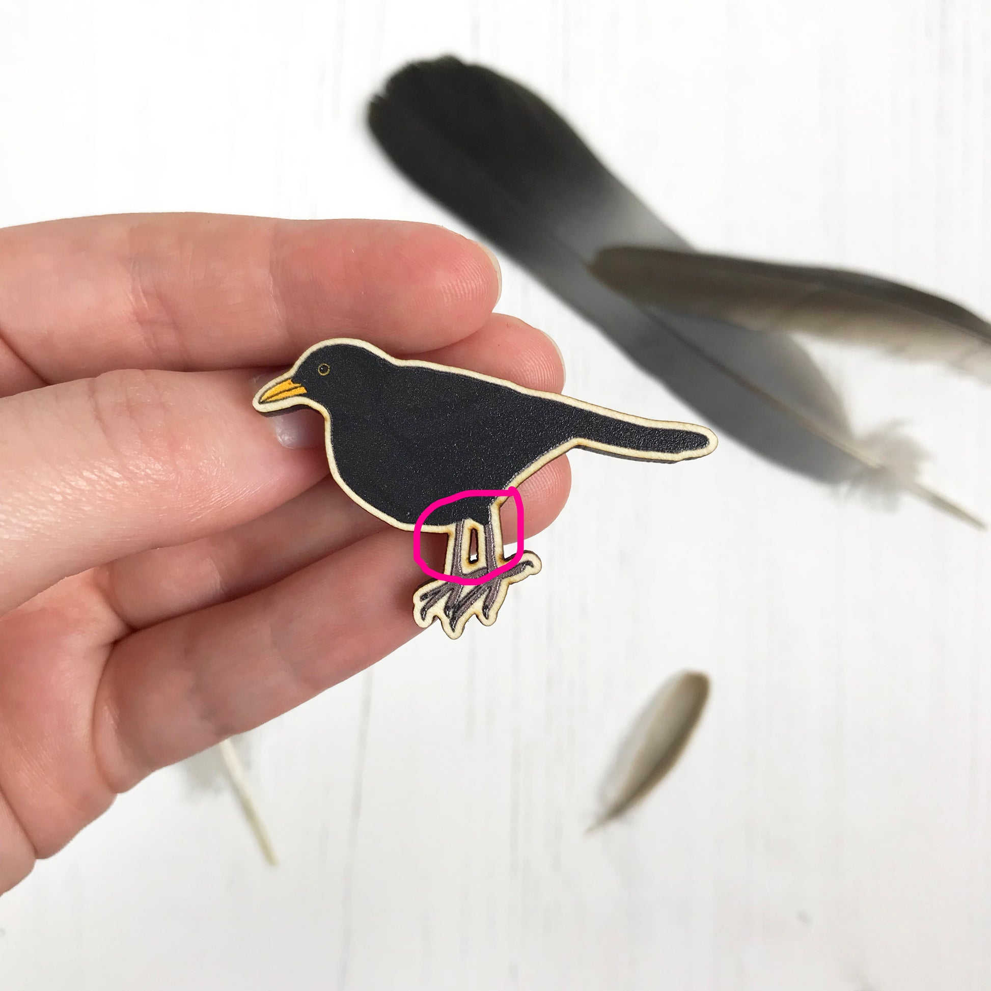 A laser cut wooden brooch showing an illustration by Loadofolbobbins of a male blackbird. A pink circle highlights some laser burns on some edges.