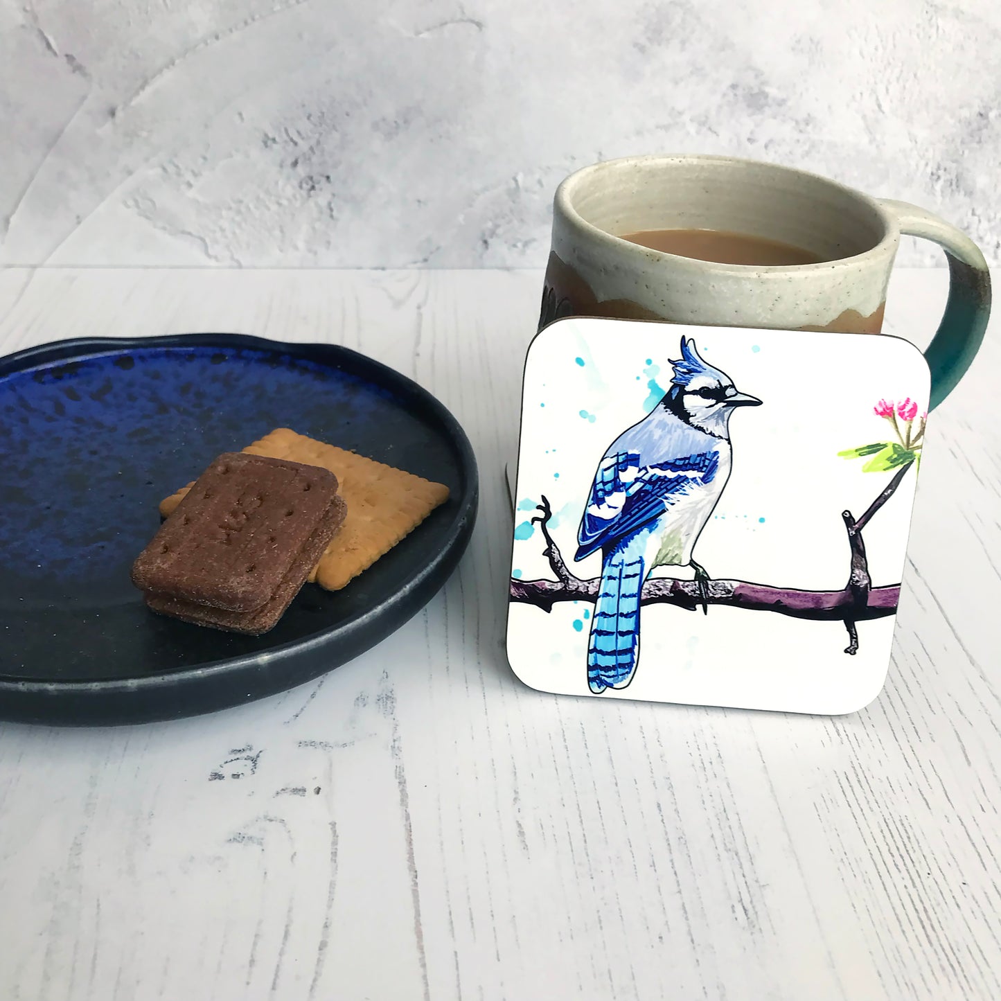 A photo of a round edged square coaster which has an original illustration by Loadofolbobbins showing  a bluejay bird perching on a reddish brown gnarled branch with blossom buds on. The bird has it’s back to the viewer, it’s head turned to the right so you can see it’s profile. It has a crest of feathers on its head that are light blue like the back feathers. Face and under body is white. The background has some light blue inky splashes.