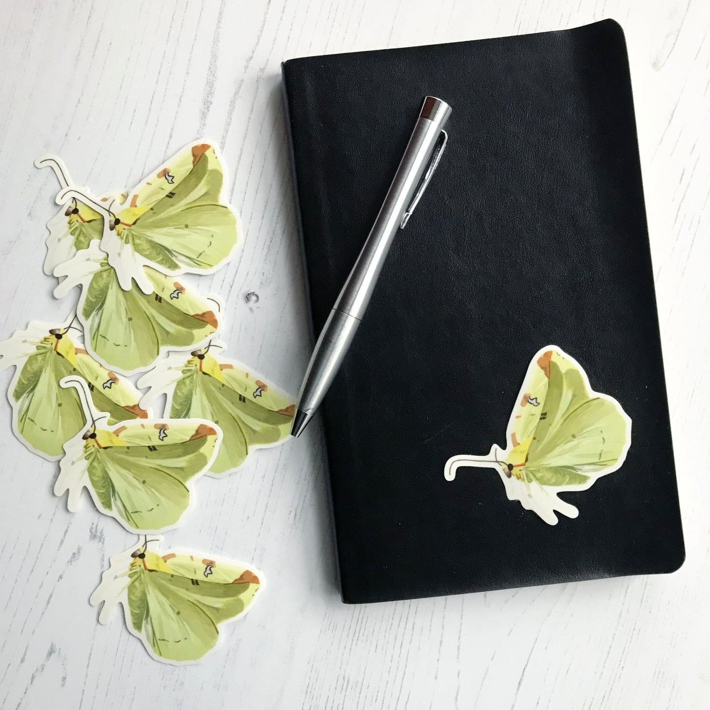 A brimstone moth sticker created from an original digital illustration by Loadofolbobbins. Shown as a side view with it's wings upright as if flapping. This moth has yellow colouring all over with light yellow wings that have an almost greenish tinge and light brown patches spaced along the top edge. The main furry section of the body has slight hints of stronger brighter yellows and it has dark compound eyes and antennae. The sticker is on a black notebook with a pen resting on it.