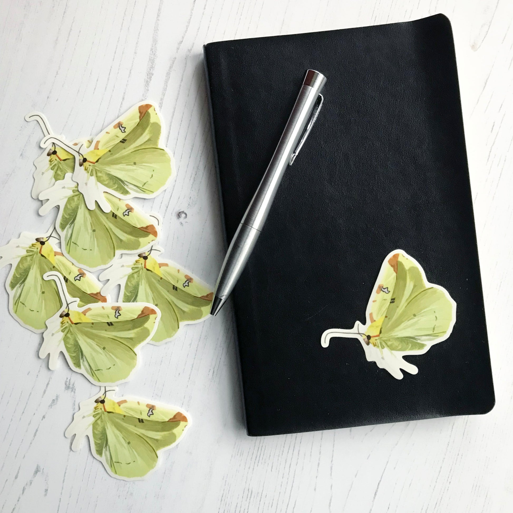 A brimstone moth sticker created from an original digital illustration by Loadofolbobbins. Shown as a side view with it's wings upright as if flapping. This moth has yellow colouring all over with light yellow wings that have an almost greenish tinge and light brown patches spaced along the top edge. The main furry section of the body has slight hints of stronger brighter yellows and it has dark compound eyes and antennae. The sticker is on a black notebook with a pen resting on it.