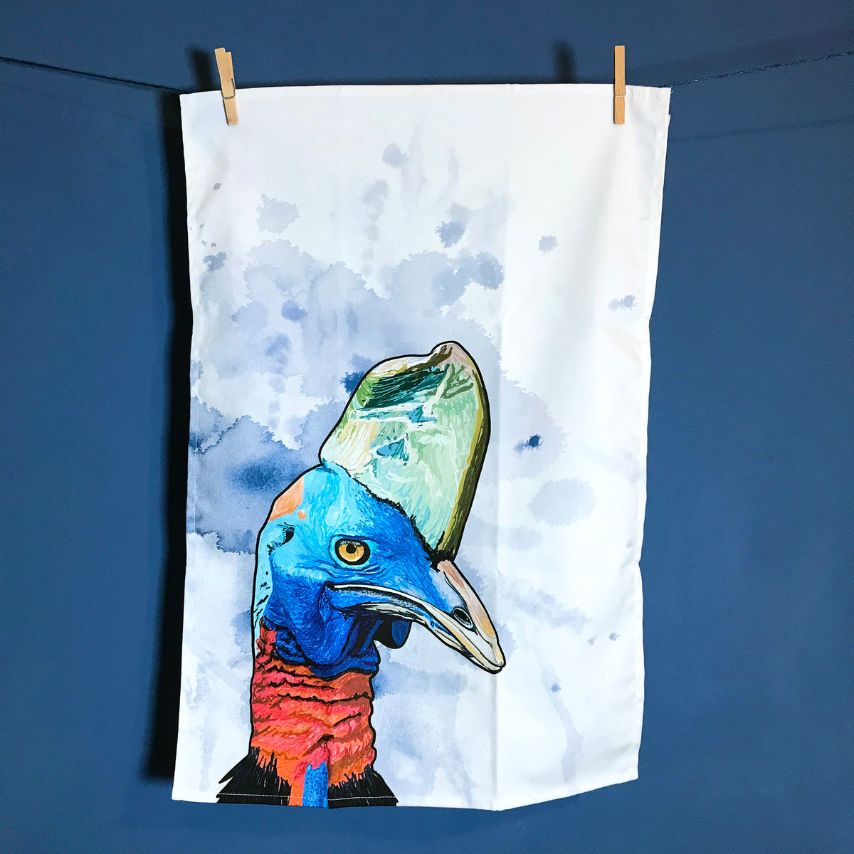 A  photo of  tea towel hanging on a washing line against a dark blue wall. The tea towel shows an illustration of the head of a Cassowary against an inky dark blue wash white background.