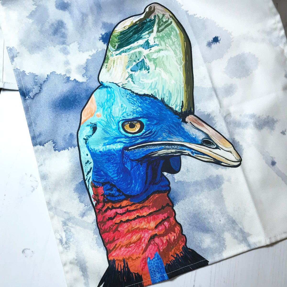 A close up photo of a tea towel showing an illustration of the head of a Cassowary against an inky wash dark blue and white background.