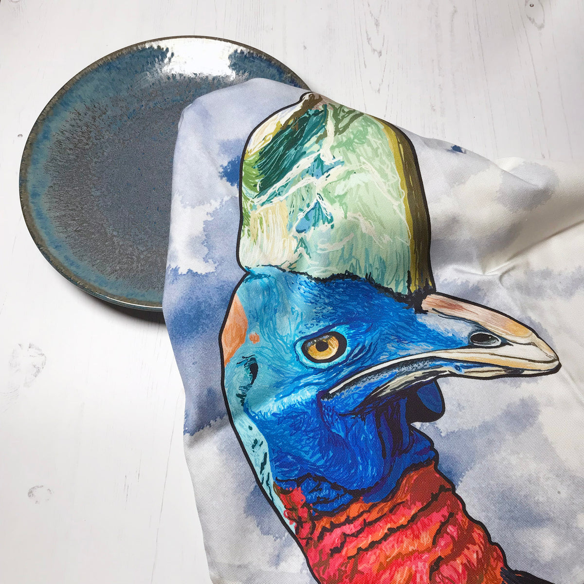 A  photo of a loosely scrunched up tea towel looking like it is drying a ceramic glazed dark turquoise plate. The tea towel shows an illustration of the head of a cassowary bird, against an inky wash dark blue and white background.