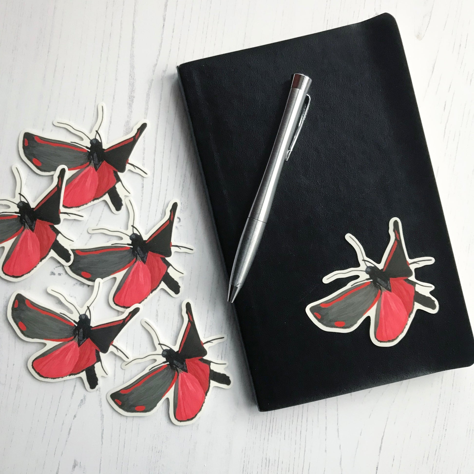 A cinnabar moth sticker created from an original digital illustration by Loadofolbobbins. The moth is mid flight with underside facing the viewer. This moth has incredibly bold markings in red and black. It's lower wings are predominantly a bright crimson with black edging. It's upper wings are deep black with crimson stripes at the top and bottom of the wings with two red dots at the far edges. It's body, legs and antennae are also a deep black. The sticker is on a black notebook with a pen resting on it.