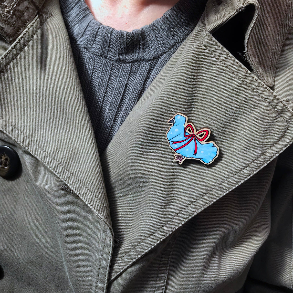 A laser cut wooden lapel pin showing an original illustration by Loadofolbobbins of a pigeon wrapped up like a Christmas present with a big red ribbon,it's beak and feet are peeking out of the wrapping paper. The style of the illustration is simplified with flat colours and a bold black outline. The pin is shown worn on the lapel of the jacket.
