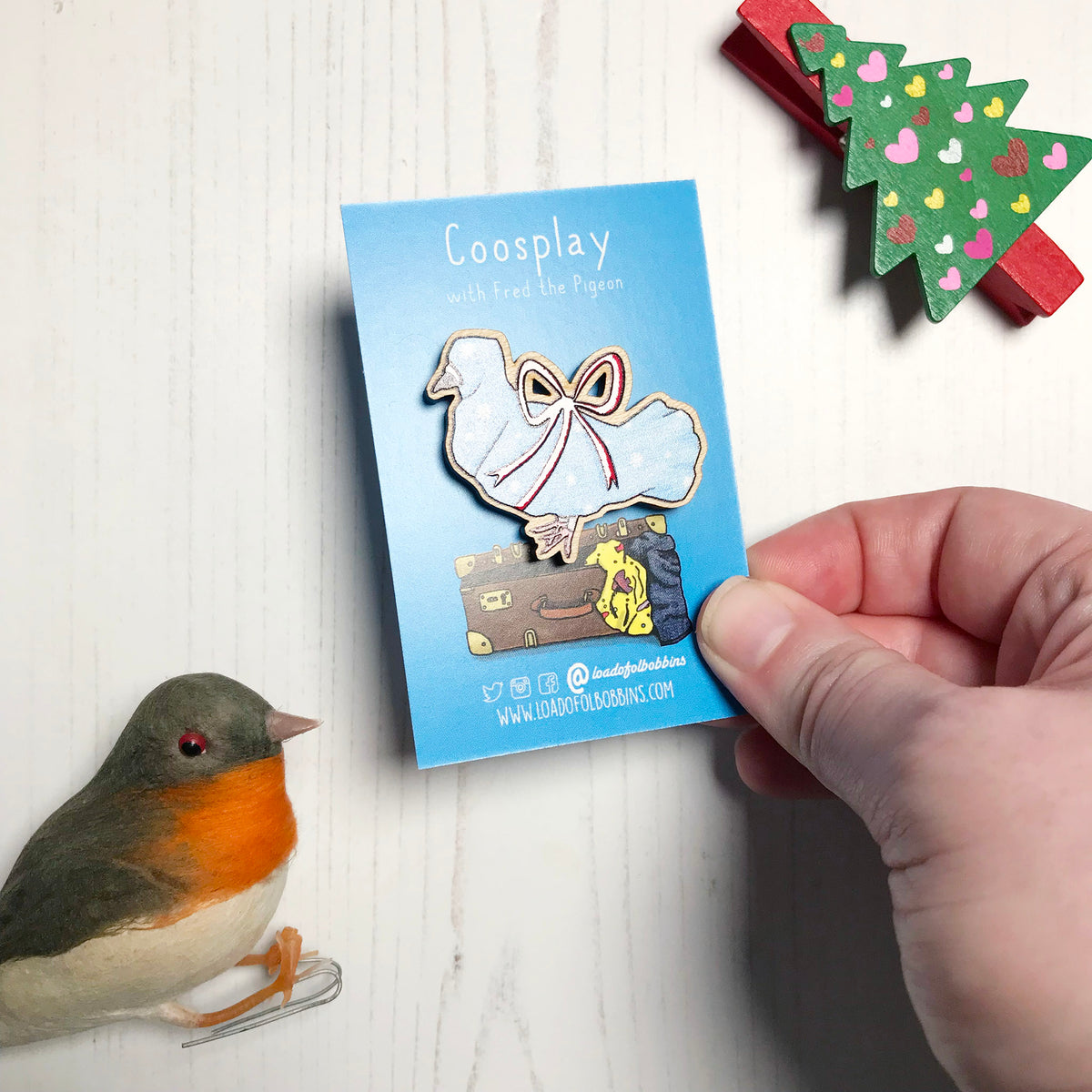 A laser cut wooden lapel pin showing an original illustration by Loadofolbobbins of a pigeon wrapped up like a Christmas present with a big red ribbon,it's beak and feet are peeking out of the wrapping paper. The style of the illustration is simplified with flat colours and a bold black outline. The pin is mounted on a blue backing card that says Coosplay with Fred the pigeon. It's held at an angle showing off the spot gloss detail on the red ribbon. 