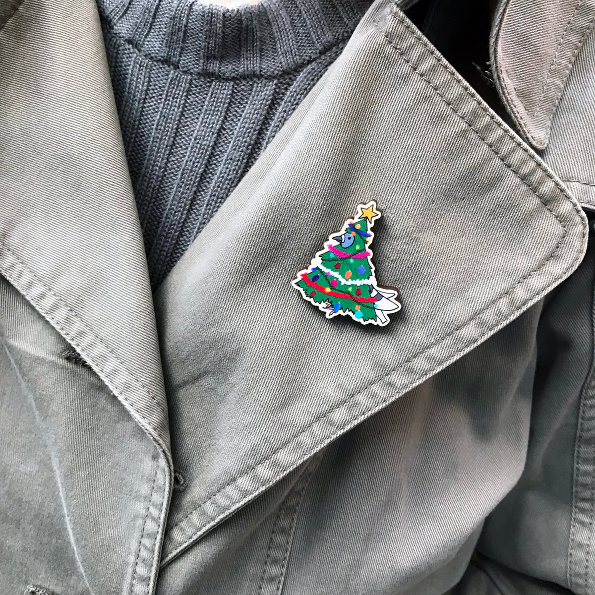 A laser cut wooden lapel pin showing an original illustration by Loadofolbobbins of a pigeon dressed as a Christmas Tree, complete with tinsel, decorations and star. The style of the illustration is simplified with flat colours and a bold black outline. The pin is shown worn on the lapel of a jacket.
