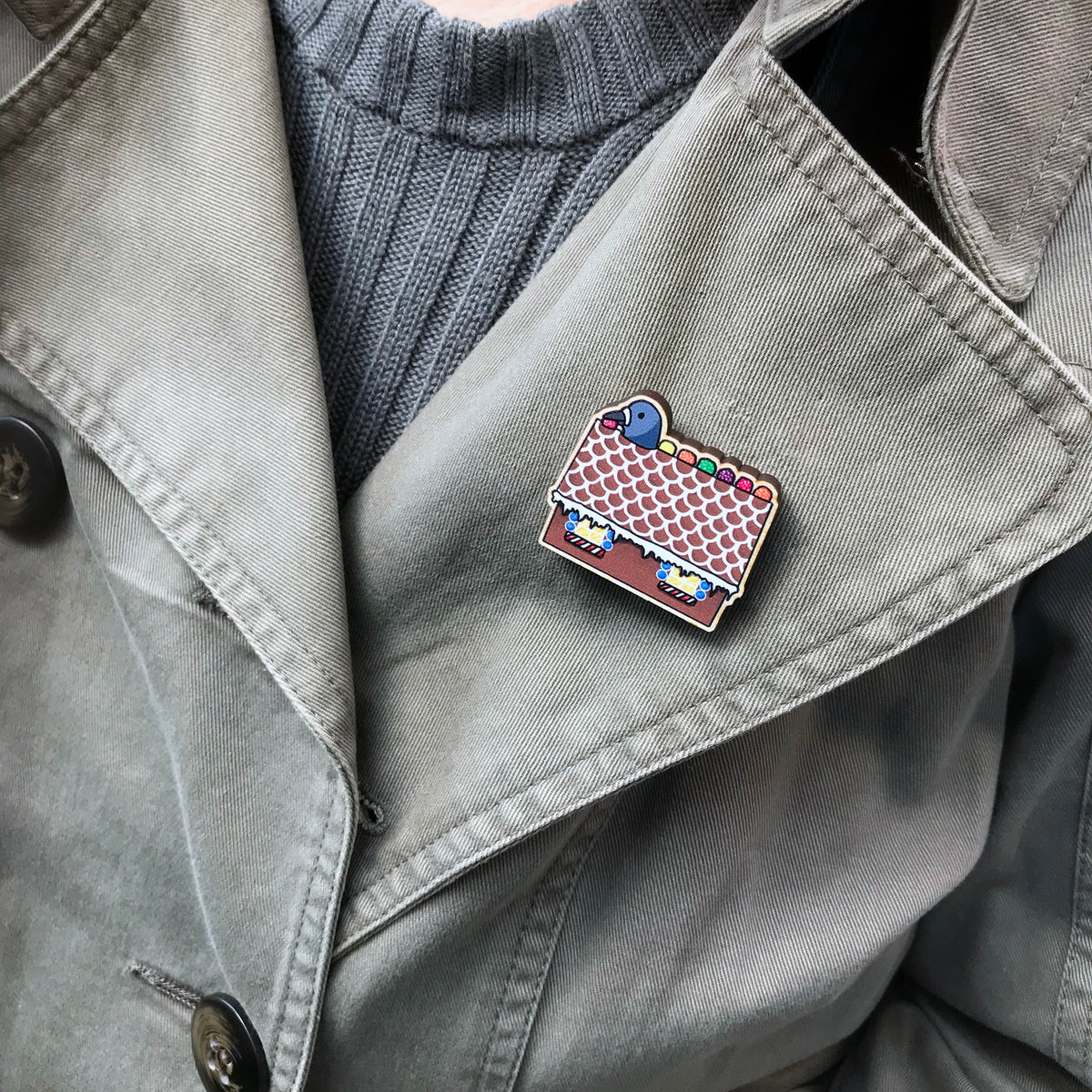 A laser cut wooden lapel pin showing an original illustration by Loadofolbobbins of a pigeon with it's head popping out of a gingerbread house nibbling on some jelly sweets.. The style of the illustration is simplified with flat colours and a bold black outline. The pin is shown worn on the lapel of a jacket.