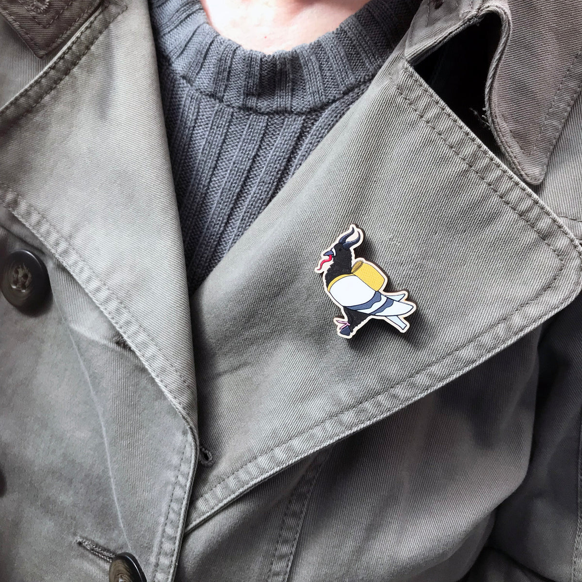 A laser cut wooden lapel pin showing an original illustration by Loadofolbobbins of a pigeon dressed as Krampus, with horns a cloven hoof and long tongue. The style of the illustration is simplified with flat colours and a bold black outline. The pin is shown on the lapel of the jacket.