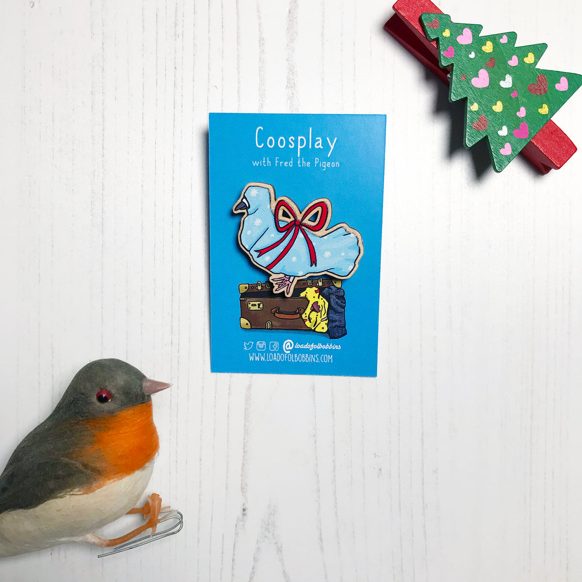 A laser cut wooden lapel pin showing an original illustration by Loadofolbobbins of a pigeon wrapped up like a Christmas present with a big red ribbon,it's beak and feet are peeking out of the wrapping paper. The style of the illustration is simplified with flat colours and a bold black outline. The pin is mounted on a blue backing card that says Coosplay with Fred the pigeon.