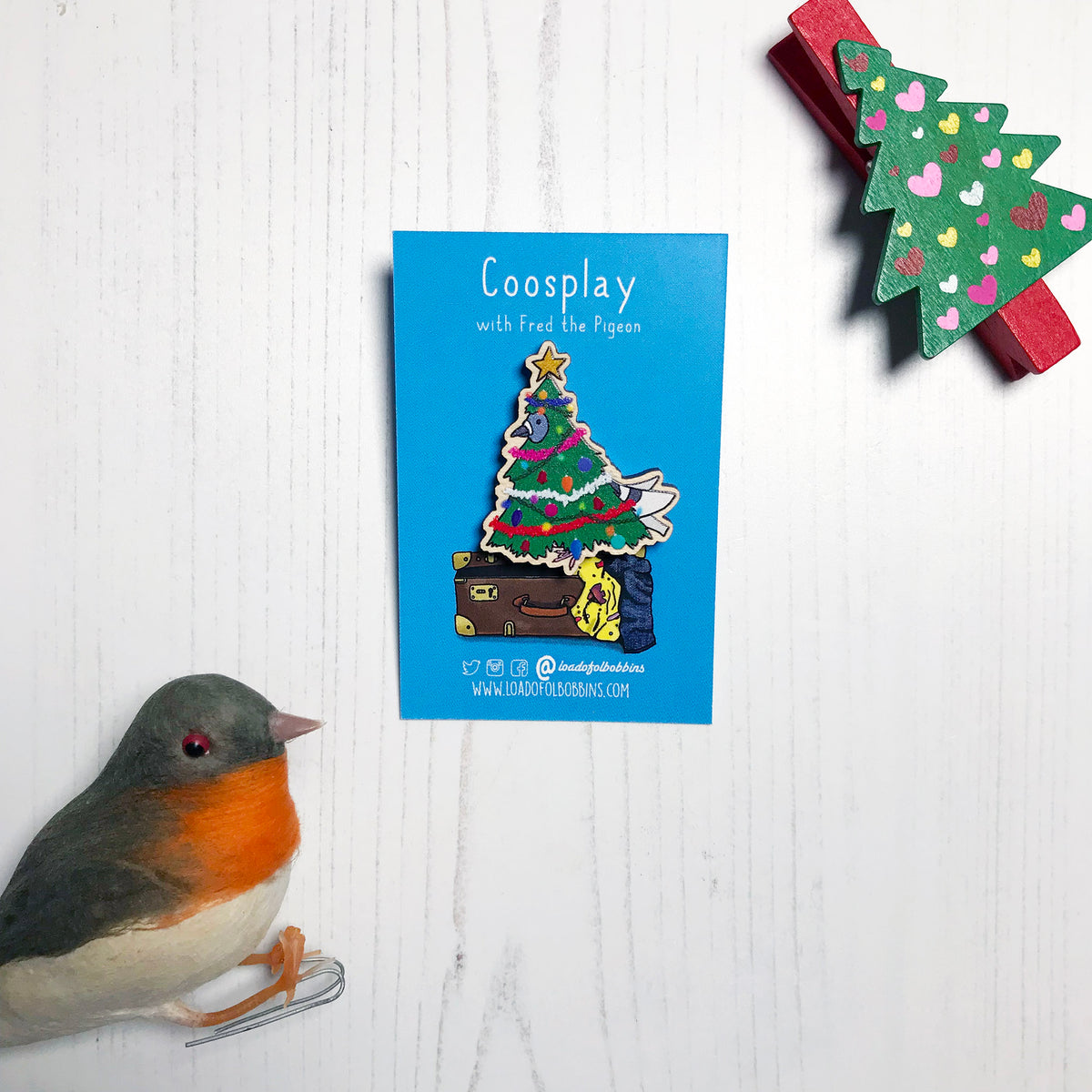 A laser cut wooden lapel pin showing an original illustration by Loadofolbobbins of a pigeon dressed as a Christmas Tree, complete with tinsel, decorations and star. The style of the illustration is simplified with flat colours and a bold black outline. The pin is mounted on a blue backing card that says Coosplay with Fred the pigeon.