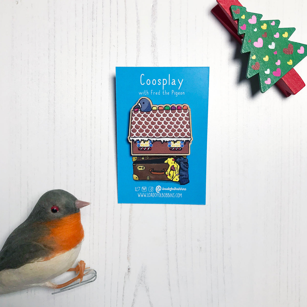 A laser cut wooden lapel pin showing an original illustration by Loadofolbobbins of a pigeon with it's head popping out of a gingerbread house nibbling on some jelly sweets.. The style of the illustration is simplified with flat colours and a bold black outline. The pin is mounted on a blue backing card that says Coosplay with Fred the pigeon.