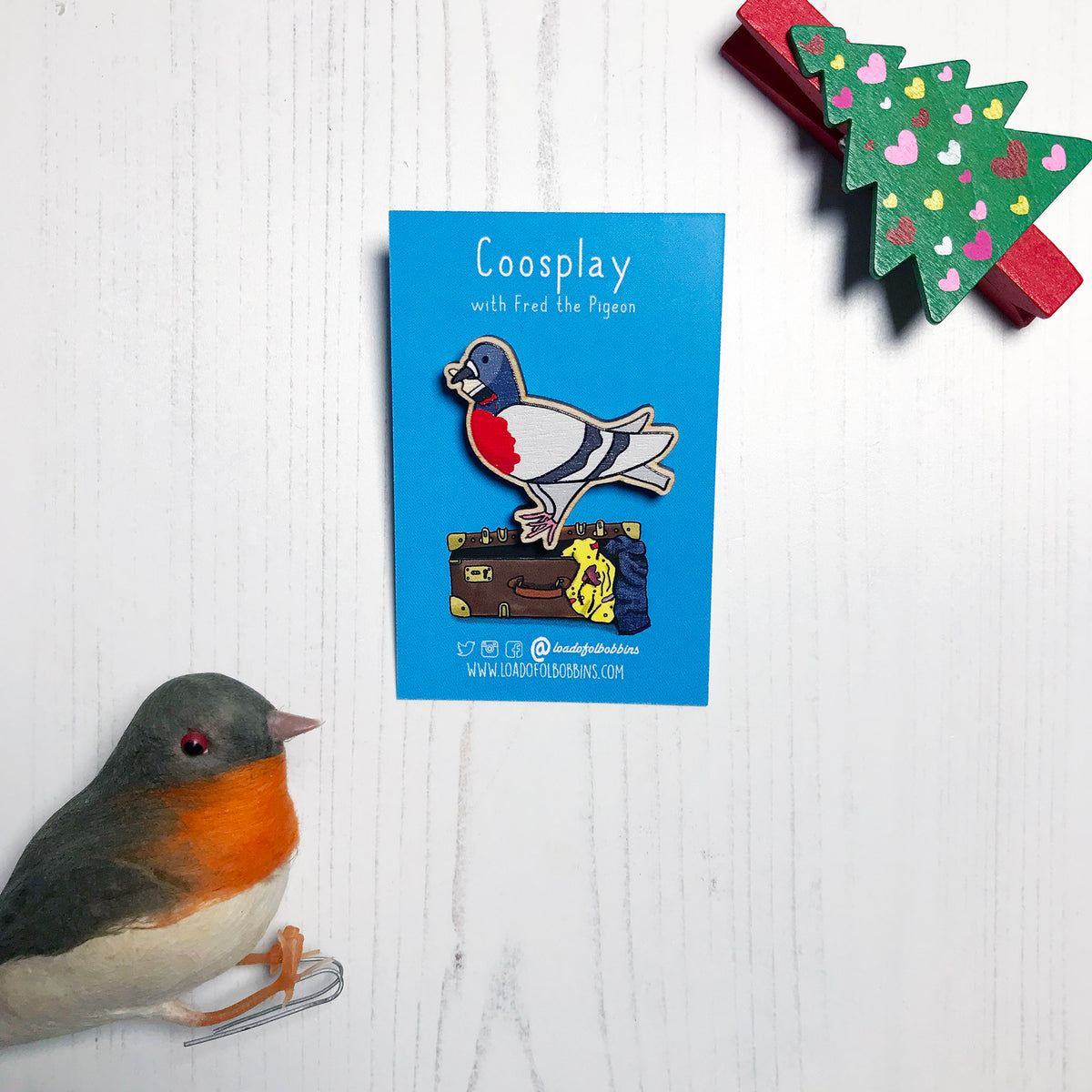 A laser cut wooden lapel pin showing an original illustration by Loadofolbobbins of a pigeon painting it's chest red to look like a robin. The style of the illustration is simplified with flat colours and a bold black outline. The pin is mounted on a blue backing card that says Coosplay with Fred the pigeon.