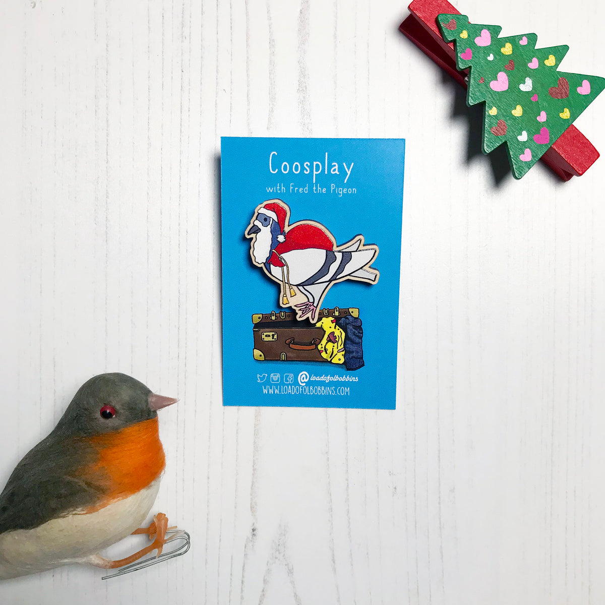 A laser cut wooden lapel pin showing an original illustration by Loadofolbobbins of a pigeon painting dressed as Santa Claus with a present sack on it's back. The style of the illustration is simplified with flat colours and a bold black outline. The pin is mounted on a blue backing card that says Coosplay with Fred the pigeon.