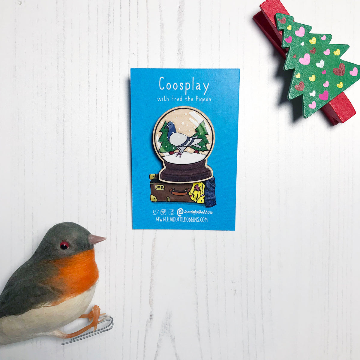 A laser cut wooden lapel pin showing an original illustration by Loadofolbobbins of a pigeon in a snow globe, standing on snow and next to two trees. The style of the illustration is simplified with flat colours and a bold black outline. The pin is mounted on a blue backing card that says Coosplay with Fred the pigeon.