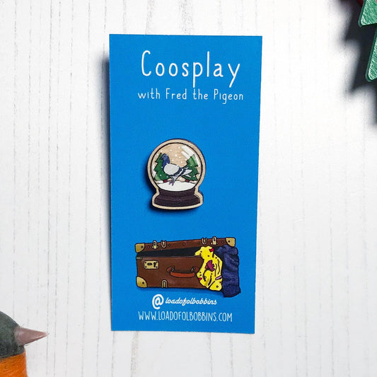 A laser cut wooden 20mm lapel pin showing an original illustration by Loadofolbobbins of a pigeon in a snow globe, standing on snow and next to two trees. The style of the illustration is simplified with flat colours and a bold black outline. The pin is mounted on a blue backing card that says Coosplay with Fred the pigeon.