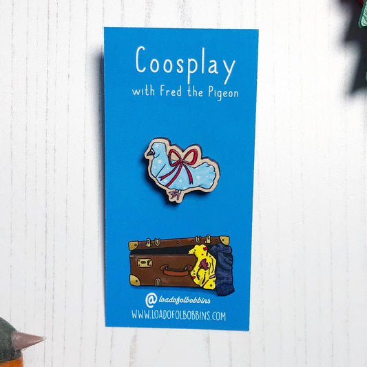 A laser cut wooden mini lapel pin showing an original illustration by Loadofolbobbins of a pigeon wrapped up like a Christmas present with a big red ribbon,it's beak and feet are peeking out of the wrapping paper. The style of the illustration is simplified with flat colours and a bold black outline. The pin is mounted on a blue backing card that says Coosplay with Fred the pigeon.
